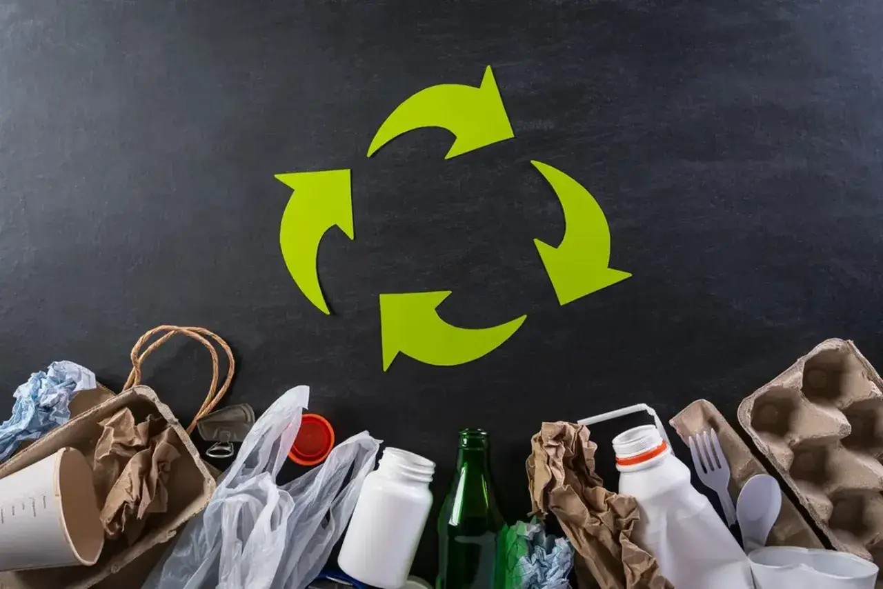 Zielony symbol recyklingu nad różnymi odpadami: papier, plastik, szkło.