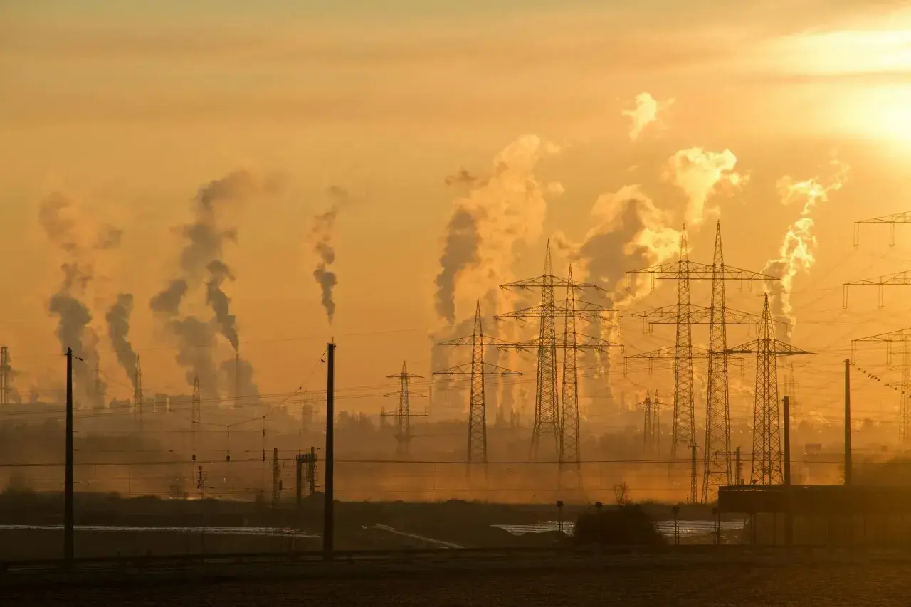 Dymiące kominy fabryk i linie energetyczne na tle zachodzącego słońca. To obraz przemysłowego krajobrazu, który ilustruje przyczyny zmian klimatu.