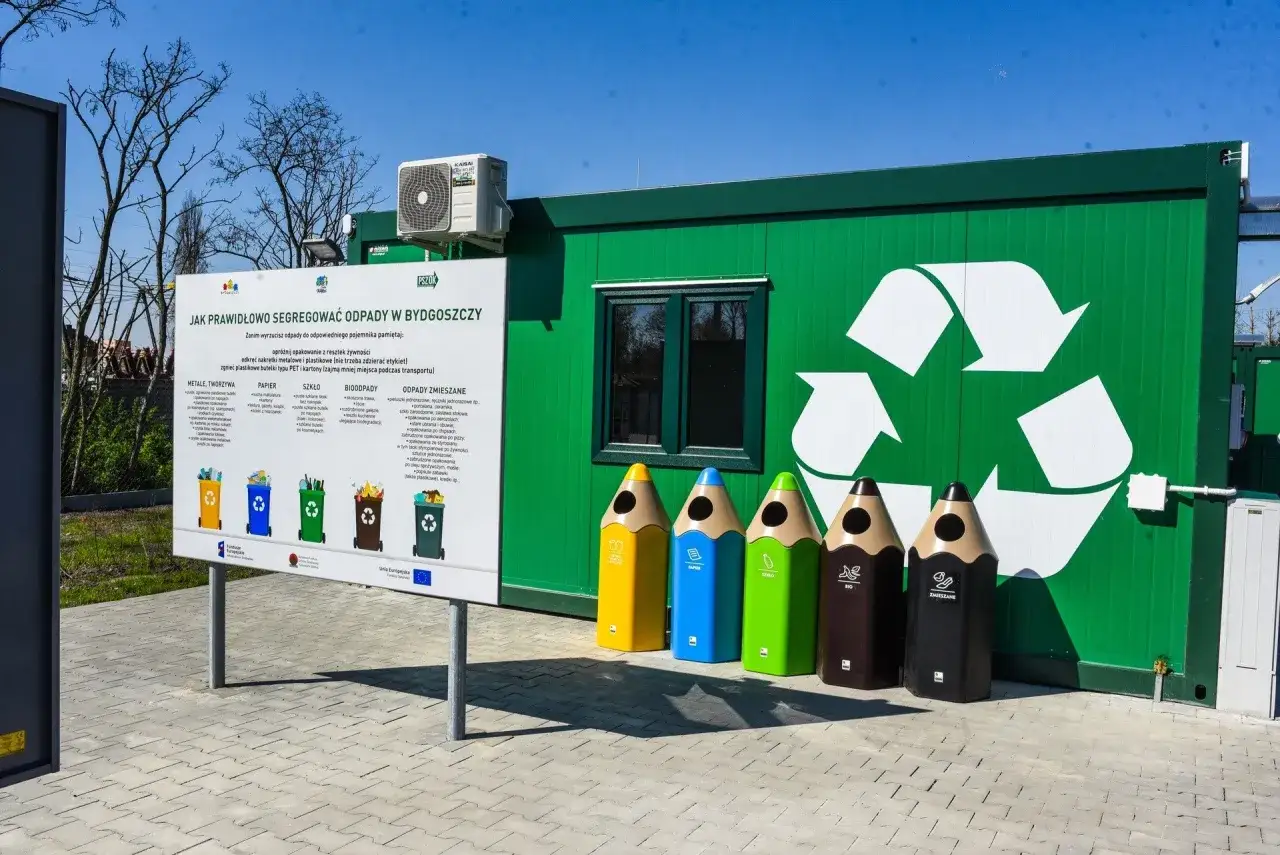 Kolorowe pojemniki w kształcie ołówków zachęcają do segregacji odpadów budowlanych. Obok tablica informacyjna i zielony kontener z symbolem recyklingu.
