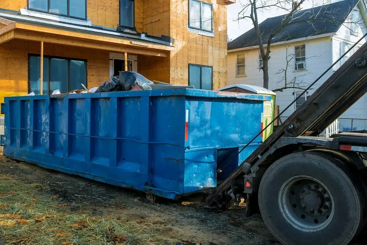 Duży, niebieski kontener na śmieci budowlane, czyli gabaryty śmieci, stoi obok domu w trakcie budowy.