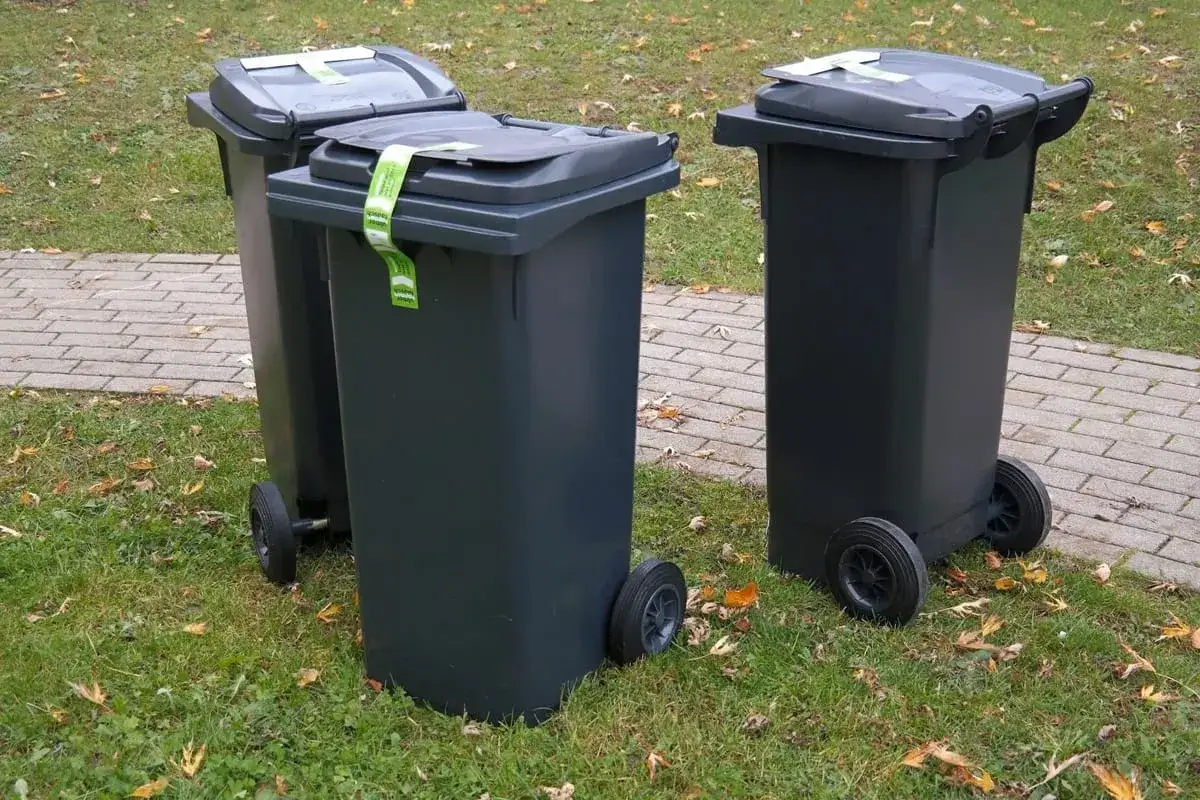 Trzy szare kosze na śmieci stoją na trawie obok chodnika. Zastanawiasz się, gdzie wyrzucić gąbkę z materaca? Sprawdź lokalne przepisy dotyczące utylizacji odpadów wielkogabarytowych.