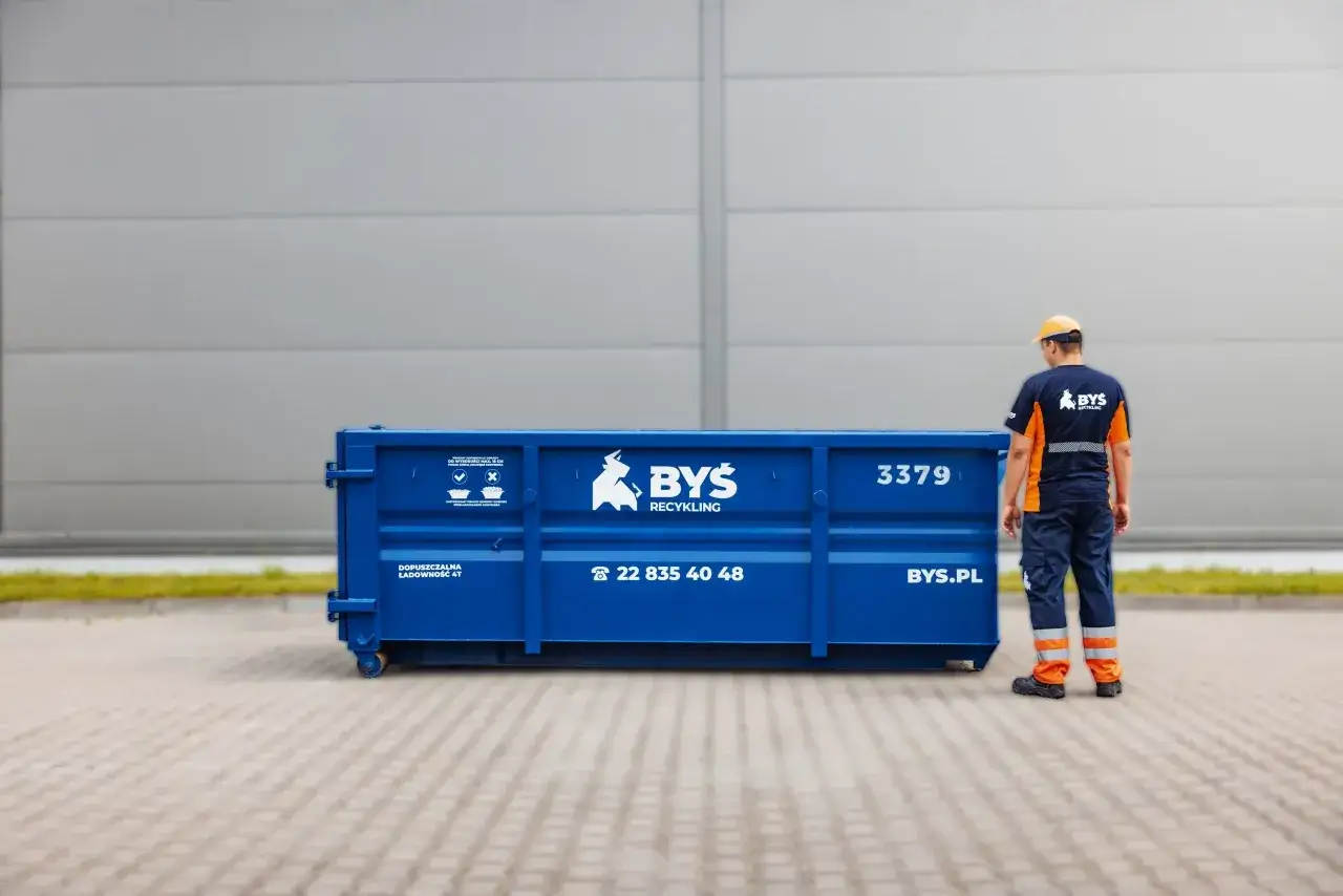 Pracownik firmy BYŚ Recycling stoi obok niebieskiego kontenera. Masz problem, gdzie wyrzucić papę? Zadzwoń pod numer 22 835 40 48.