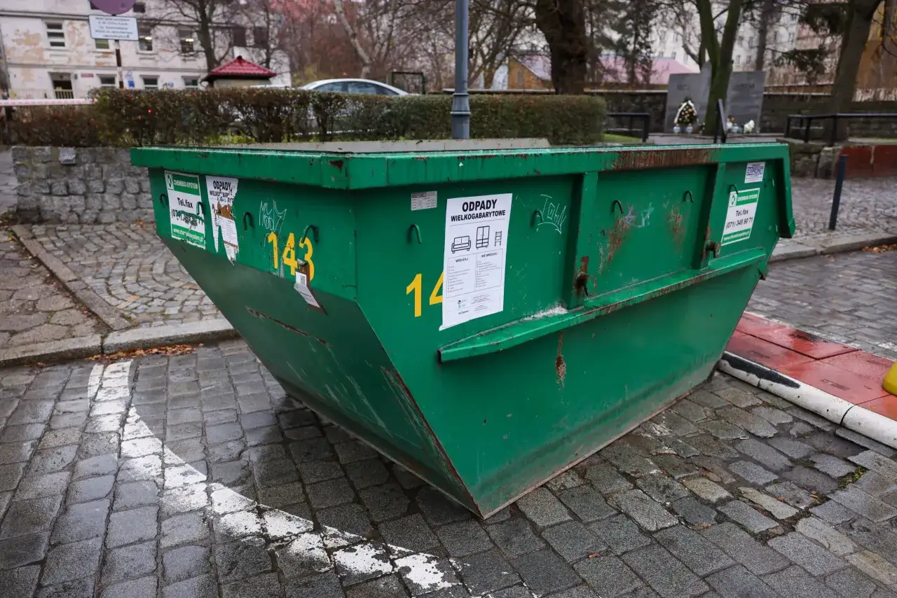 Zielony kontener na odpady wielkogabarytowe. Na nim naklejki z informacją, gdzie wyrzucić szyby okienne i inne odpady.